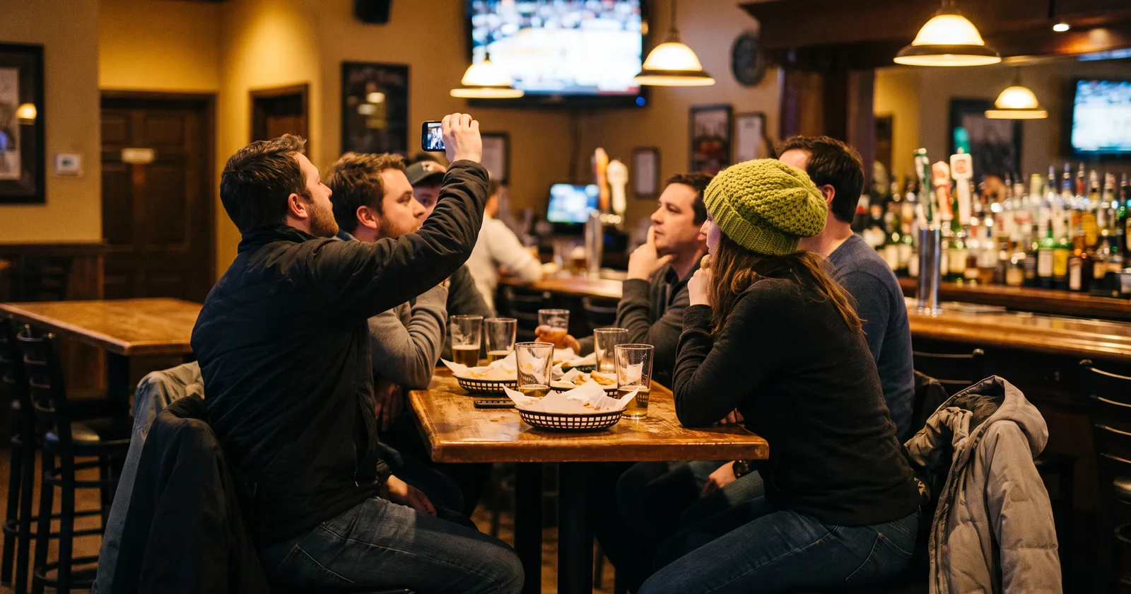 Fans at a neighbourhood sports bar on match night, seen mostly from behind, one raising a phone to check in while the group watches the game on the TV above the bar